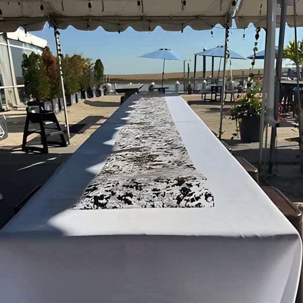 Long white table with a textured runner under a tent, with outdoor seating and blue sky in the background.