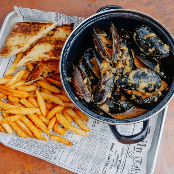 A black pot of Dox mussels with garlic sauce, fries, and toasted bread on a metal tray.