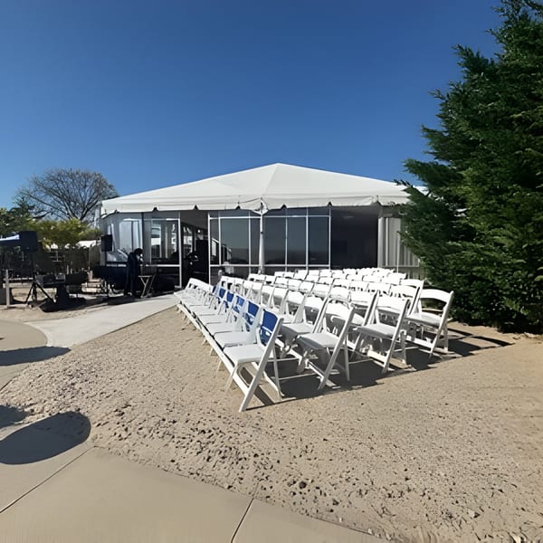 Rows of white chairs face a large white event tent on a gravel and concrete patio.